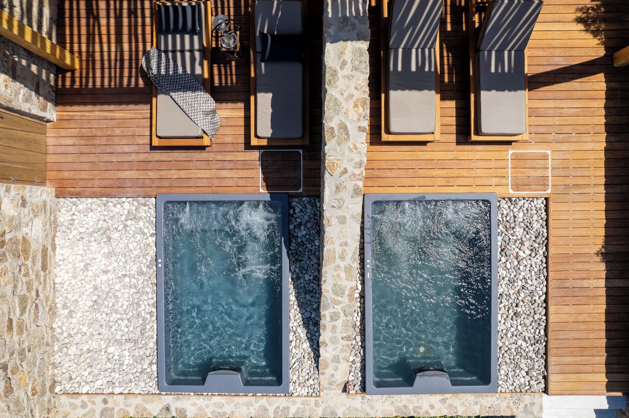 A wooden building with two hot tubs, a stone wall, and a wooden deck with lounge chairs.