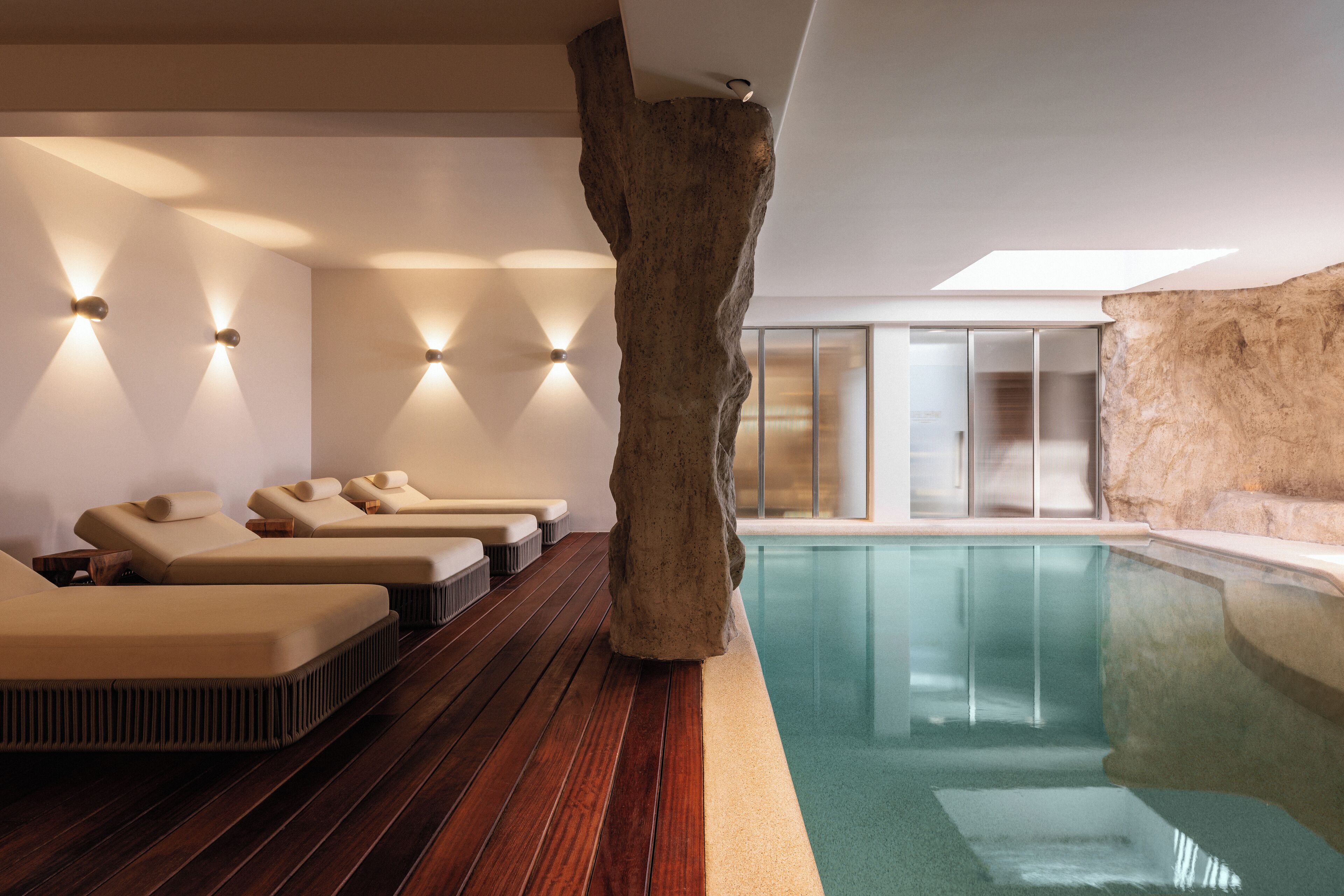 A spa area with wooden flooring, beige loungers, a large rock centerpiece, and a glass-enclosed pool.