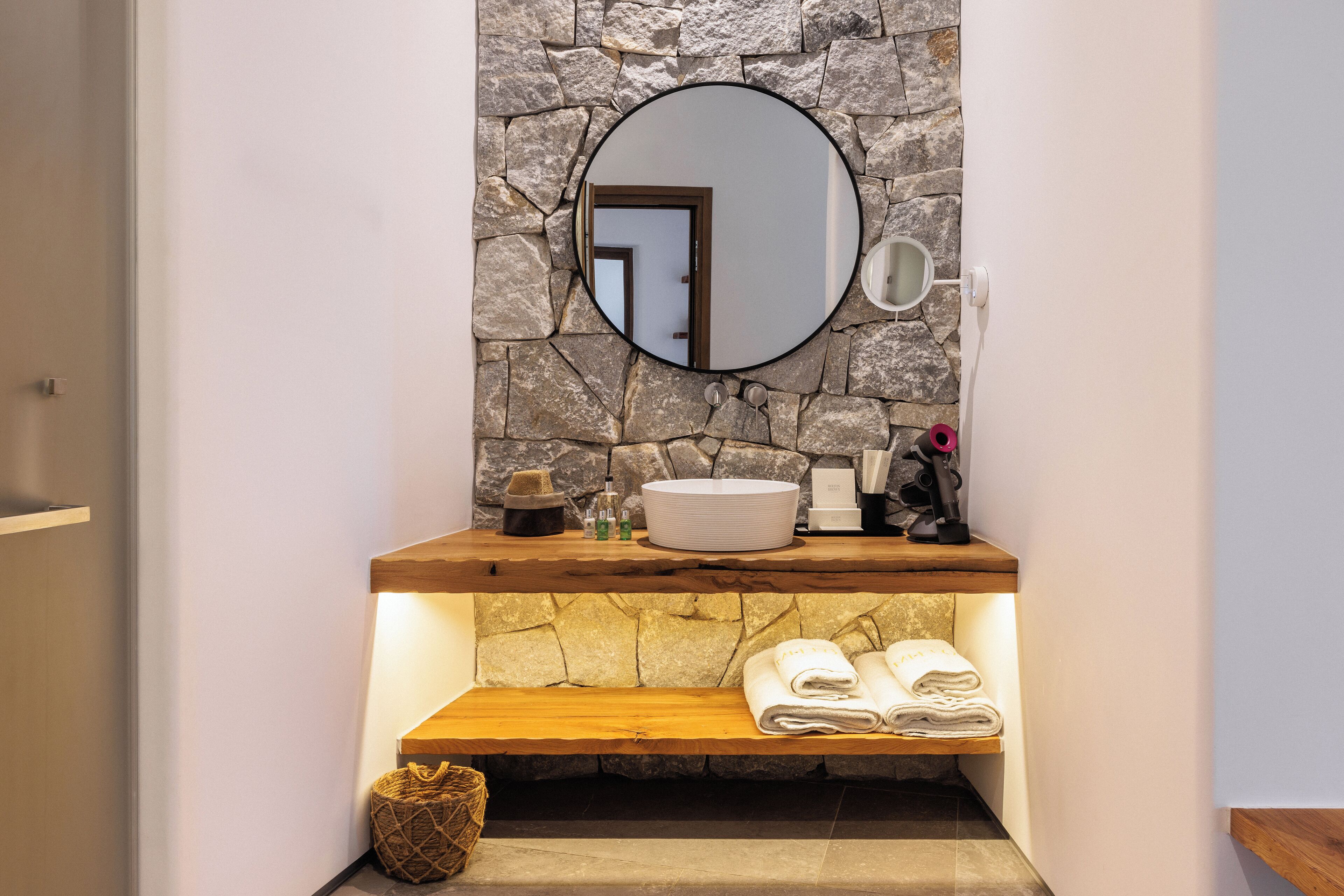 A bathroom with a stone wall, a round mirror, a wooden shelf with towels, and toiletries.