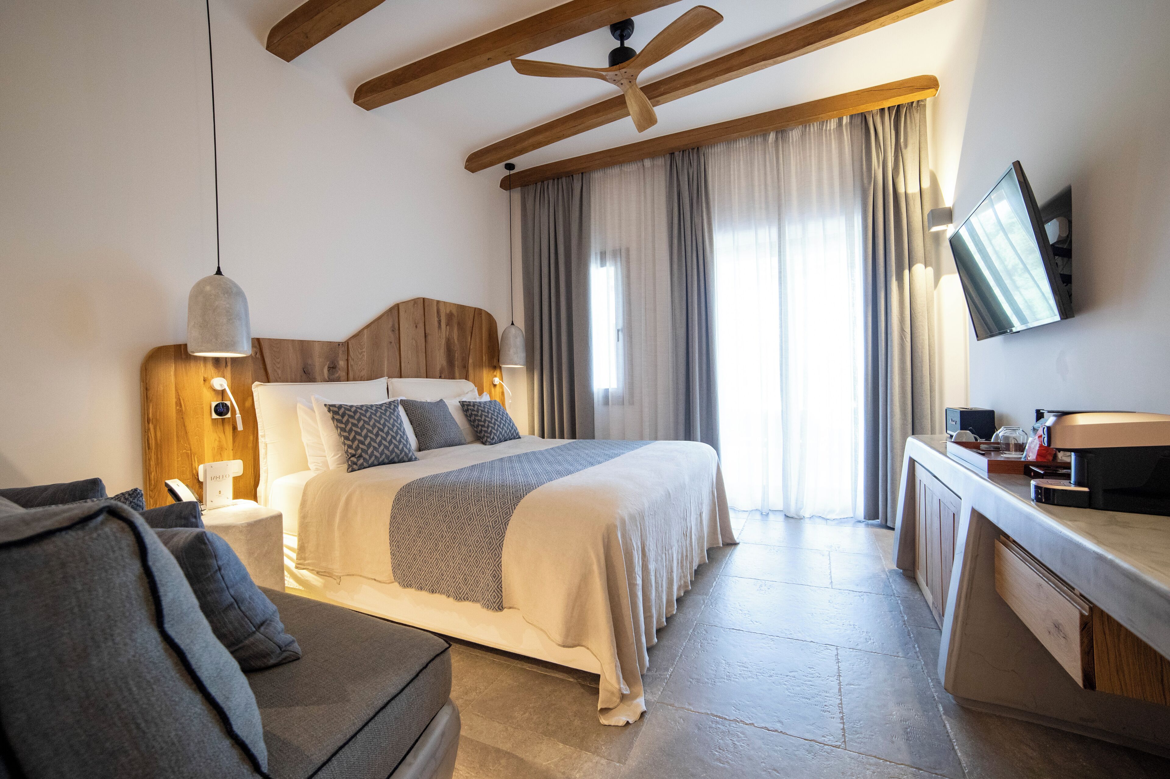 A modern hotel room with a large bed, a desk with a flat-screen TV, a ceiling fan, and a large window with curtains.