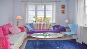 Quarto com sofá listrado, mesa redonda pequena, cadeira azul e janela com cortinas.