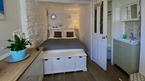 A compact bedroom with a bed, bedside table, and a small bench. A white door leads to a bathroom with a sink and cabinets. - Charming 3-bed House, Saint Ives, nr Beach & Town (St Ives)