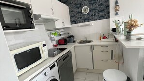 A compact kitchen with a microwave, toaster, and washing machine. There is a sink, a small trash bin, and a clock on the wall.