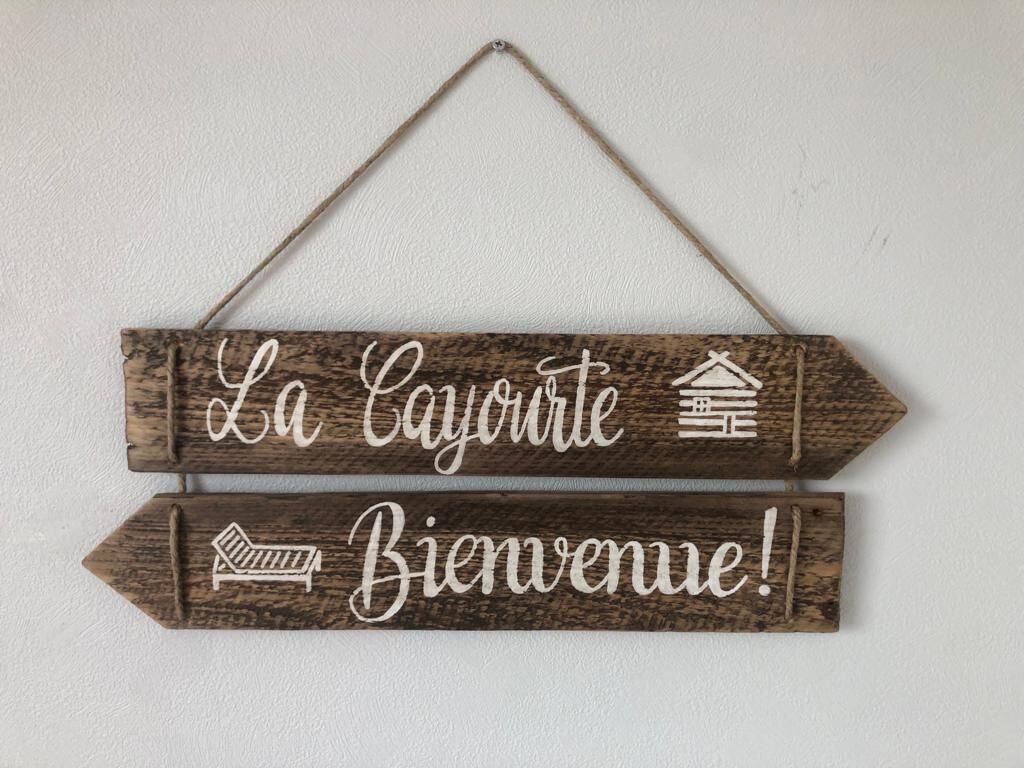 Un letrero de madera con la leyenda "La Cayenne" y "Bienvenue!", con un icono de casa y un icono de cama.
