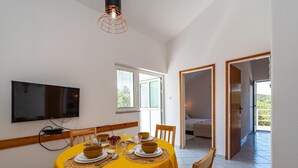 Een eetgedeelte met een gele tafel voor vier personen, een flatscreen televisie aan de wand en een raam met zonwering.