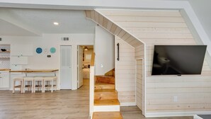 Ein modernes Interieur mit einer Holztreppe, die zu einer Schlafgalerie führt, einem Essbereich mit weißem Tisch und Stühlen sowie einem an der Wand befestigten Flachbildfernseher.