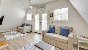 Ein geräumiges Wohnzimmer mit hoher Decke, einem Deckenventilator, einem beige farbenen Sofa mit blauen Kissen, einem weißen Couchtisch und einem weißen Beistelltisch.
