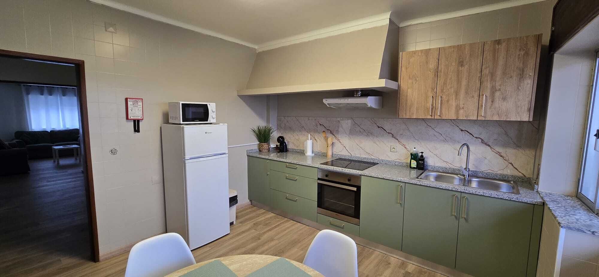 Cocina compacta con microondas, fregadero y armarios verdes. Hay un comedor con mesa y sillas.
