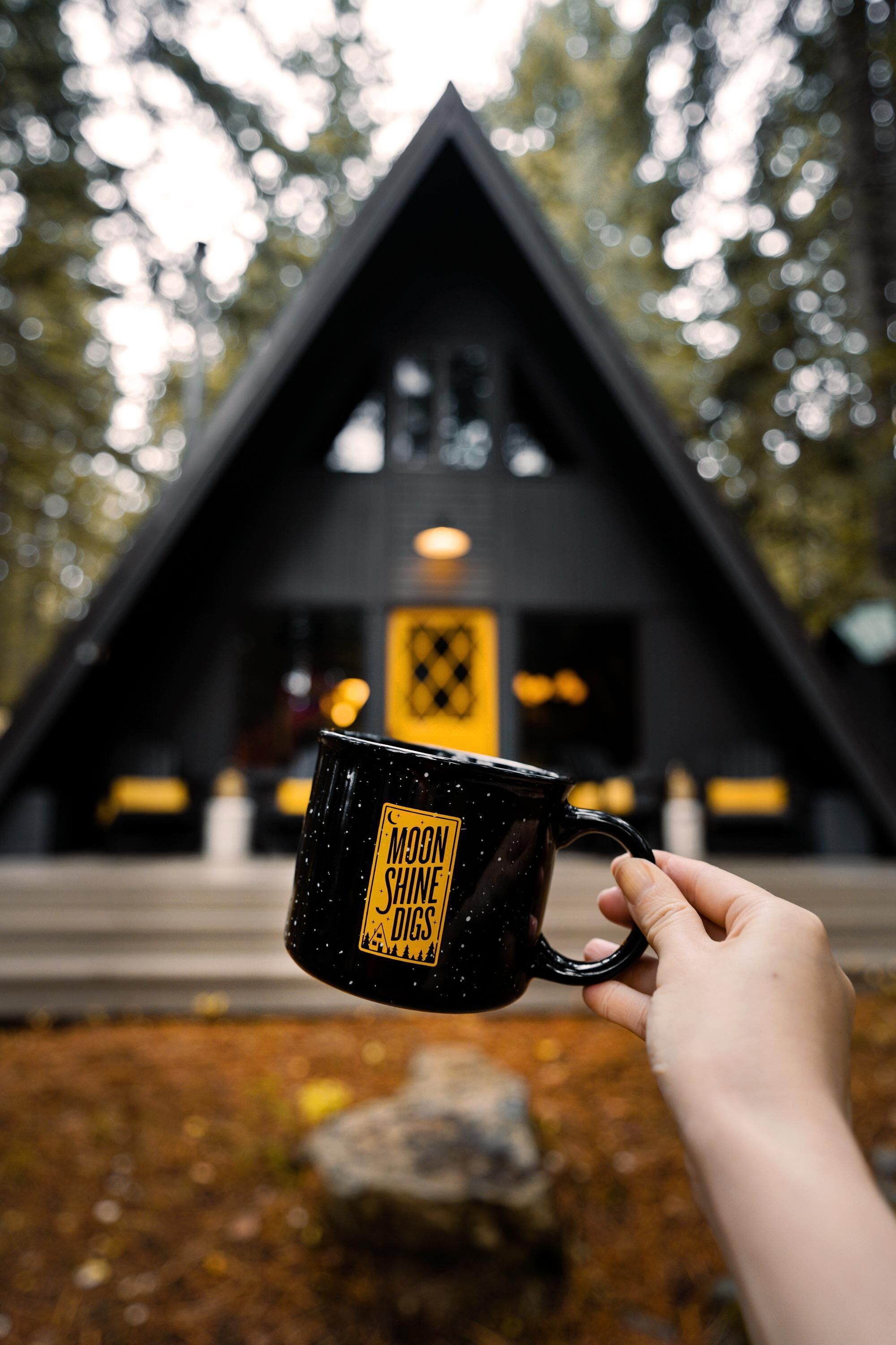 Eine Hand hält eine schwarze Tasse mit Schriftzug vor einem Gebäude mit einem dreieckigen Dach und einer beleuchteten Schrifttafel.