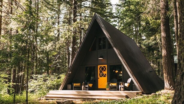 Eine dreieckige Holzhütte mit Veranda, umgeben von einem Wald.