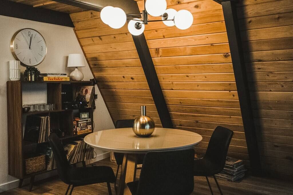 A cozy room with a wooden sloped ceiling, a round table with chairs, a clock, and a bookshelf with records.
