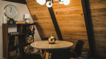 A cozy room with a wooden sloped ceiling, a round table with chairs, a clock, and a bookshelf with records.
