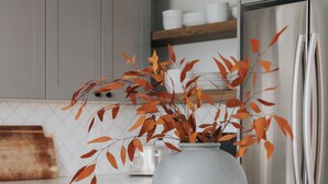 Een keuken met witte kastjes, een roestvrijstalen koelkast, een vaas met herfstbladeren en een hanglamp.