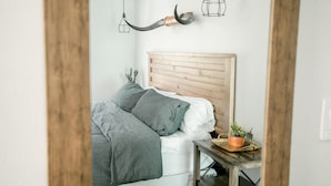 Een slaapkamer met een houten hoofdbord, een bed met grijze beddengoed, een nachtkastje met een plant en een hangende lamp.