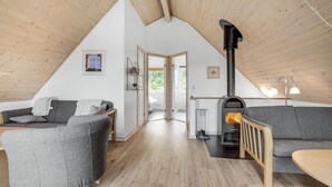 Een knus loft-achtig vertrek met een hoge plafond, houten balken, een grijze bank, een kleine tafel en een houtkachel.