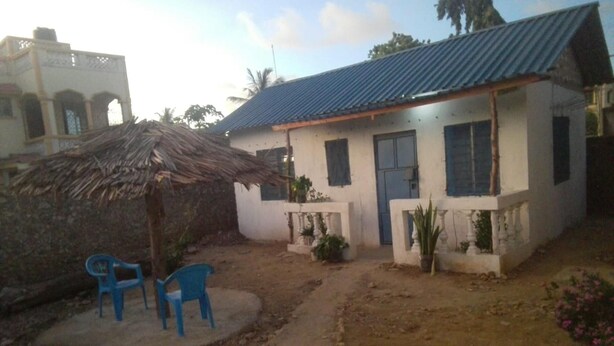 Une maison avec un toit bleu, une structure de toit en chaume et un petit jardin agrémenté de plantes en pot et d’une chaise en plastique bleue.