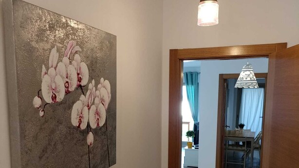 Un couloir avec, sur la gauche, un grand tableau abstrait, sur la droite, un cadre de porte en bois, et une vue sur une pièce agrémentée d’un lustre.