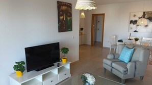Un salon moderne avec un téléviseur à écran plat posé sur un meuble blanc, une table basse en verre, un canapé gris agrémenté d’un coussin bleu, et un coin repas à l’arrière-plan.