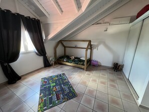Una stanza con soffitto spiovente, un letto a castello in legno, un tappetino da gioco colorato e un tavolino con una lampada.
