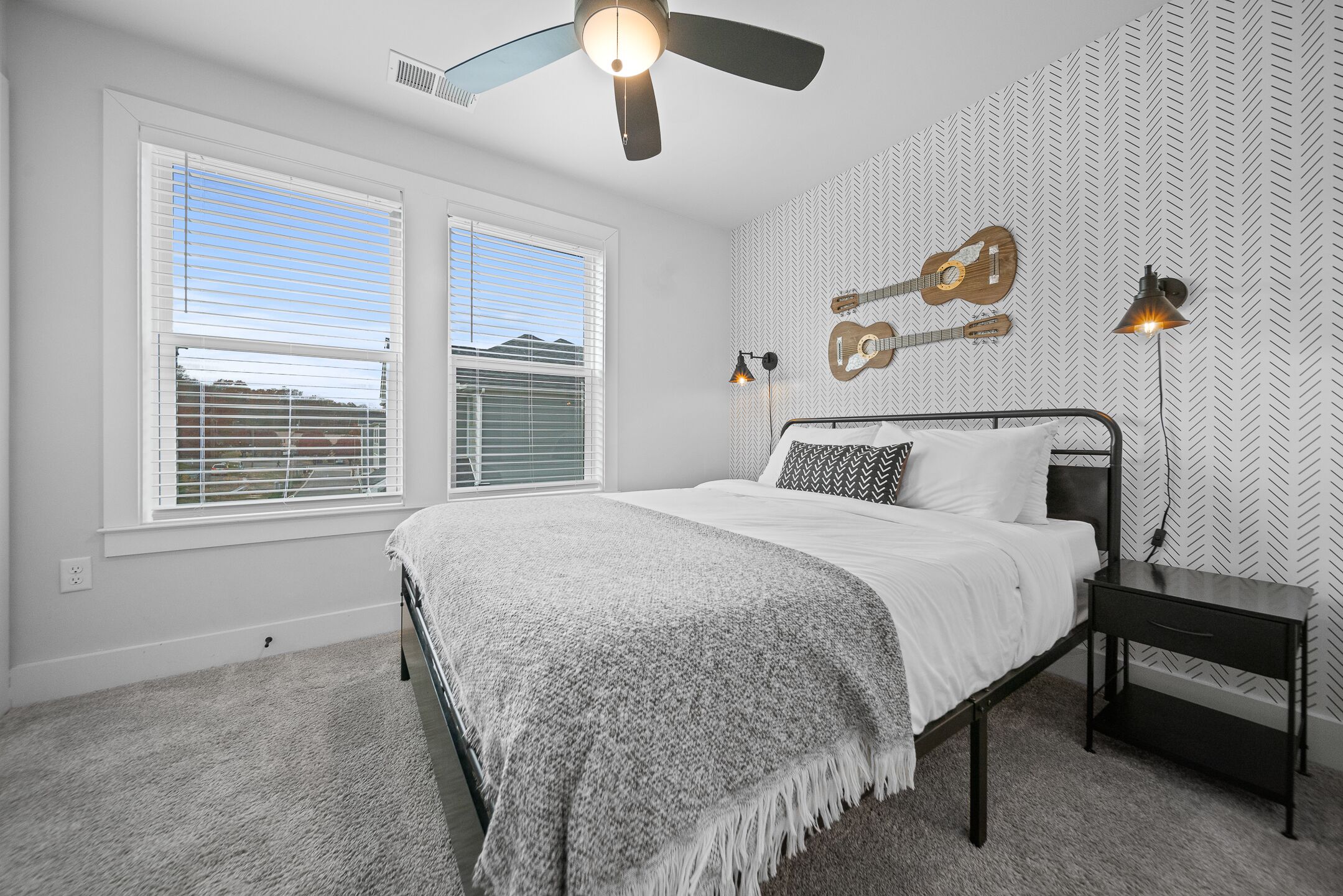 A bedroom with a bed, two windows, a ceiling fan, wall-mounted guitars, and a nightstand with a lamp.