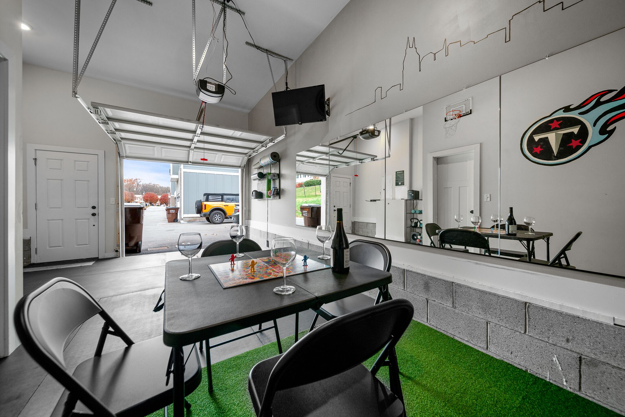 A modern indoor space with a dining table, chairs, a TV mounted on the wall, and a garage door leading outside.