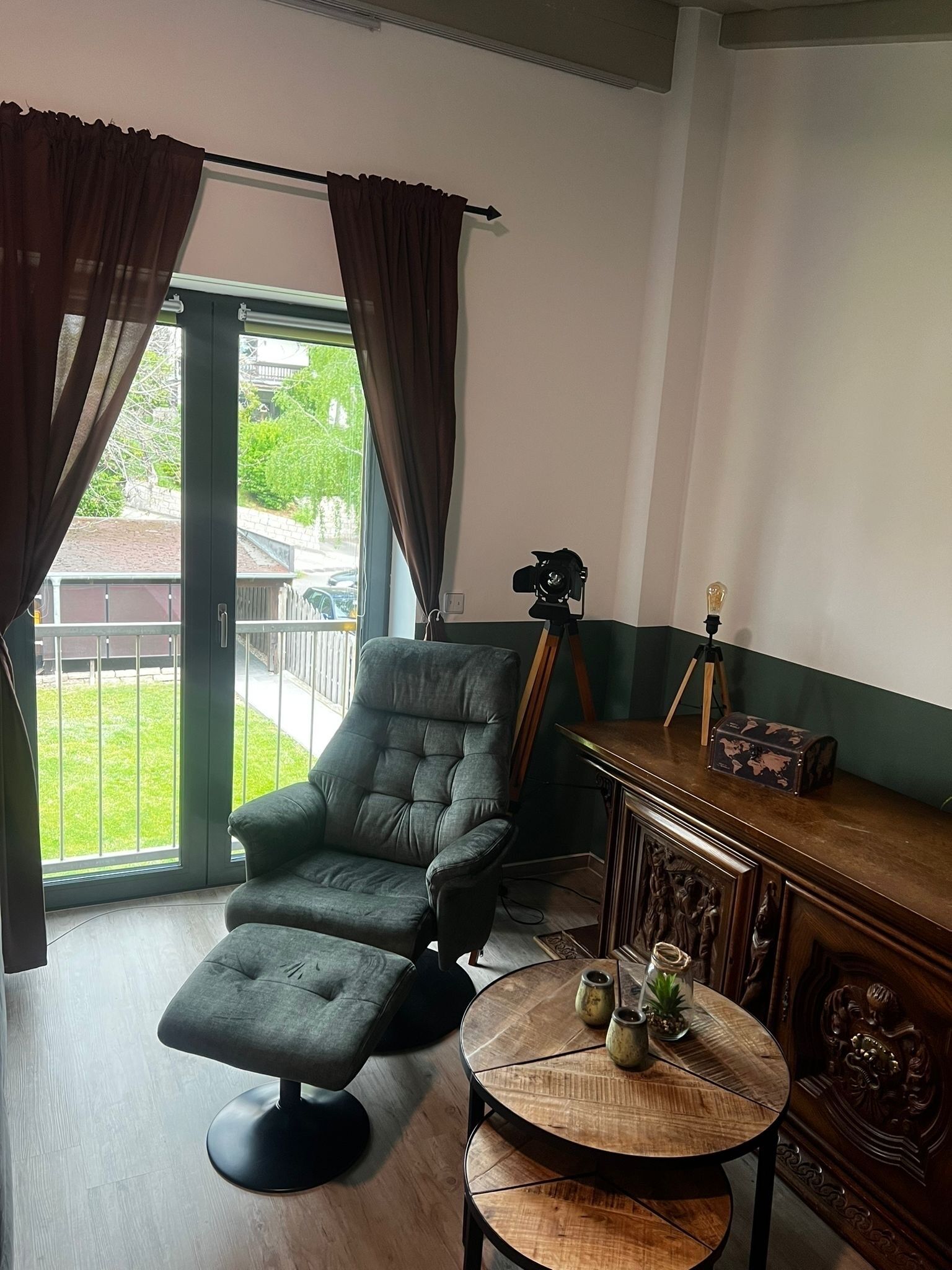 A room with a grey armchair and ottoman, a wooden side table with a world map, and a view of a green lawn and parked cars through a glass door.
