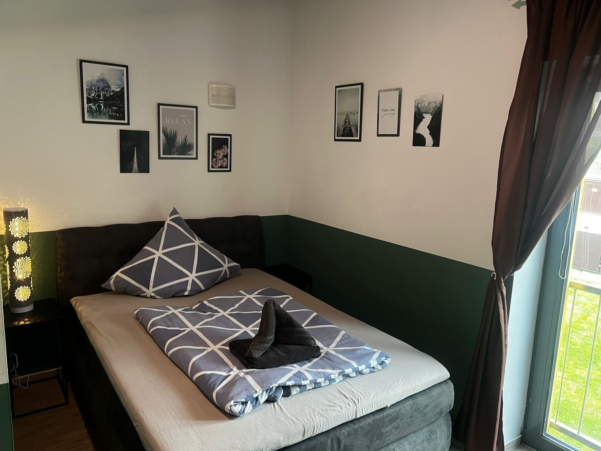 Een slaapkamer met een bed, kussens, een nachtkastje, een lamp en wanddecoratie.
