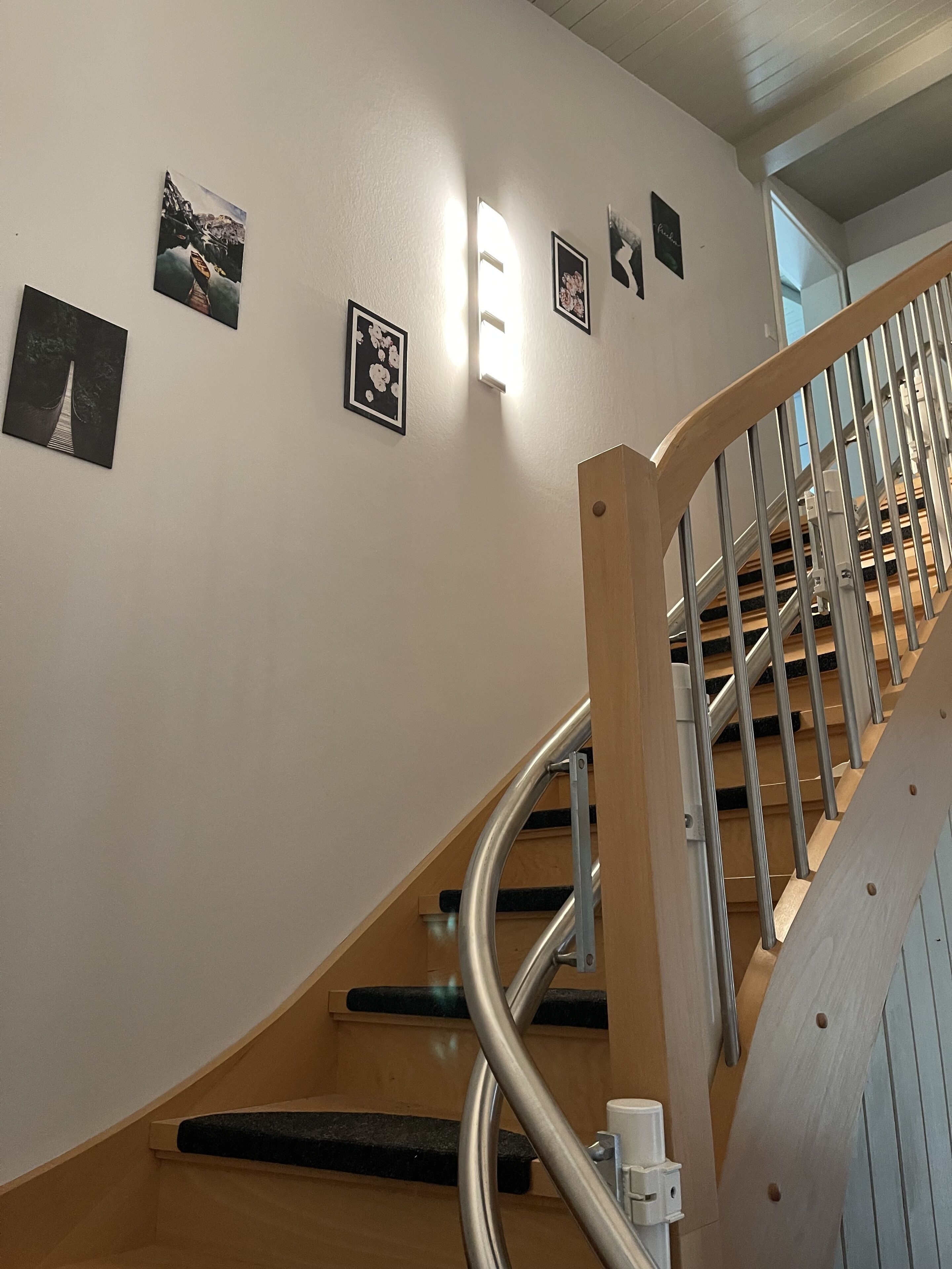 Una escalera con barandilla de madera y balaustres de metal, que conduce a un rellano con una lámpara y cuadros en la pared.