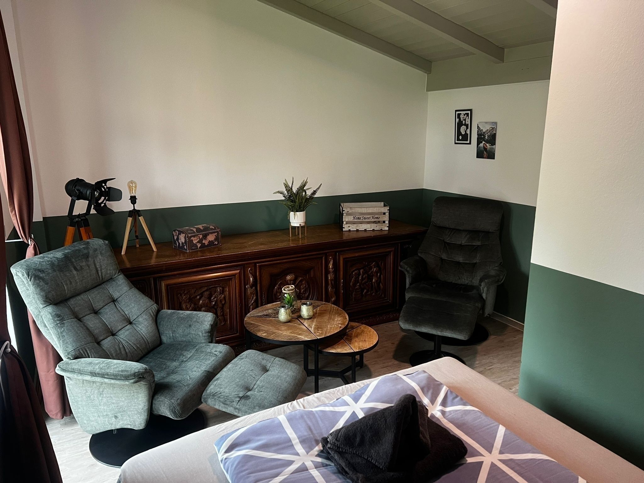 Um quarto com uma grande poltrona cinza, outra poltrona cinza menor, uma mesa de centro de madeira, e um sofá cinza escuro. Há um armário de madeira com detalhes entalhados e uma pequena planta em vaso em cima.