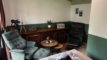 Um quarto com uma grande poltrona cinza, outra poltrona cinza menor, uma mesa de centro de madeira, e um sofá cinza escuro. Há um armário de madeira com detalhes entalhados e uma pequena planta em vaso em cima.