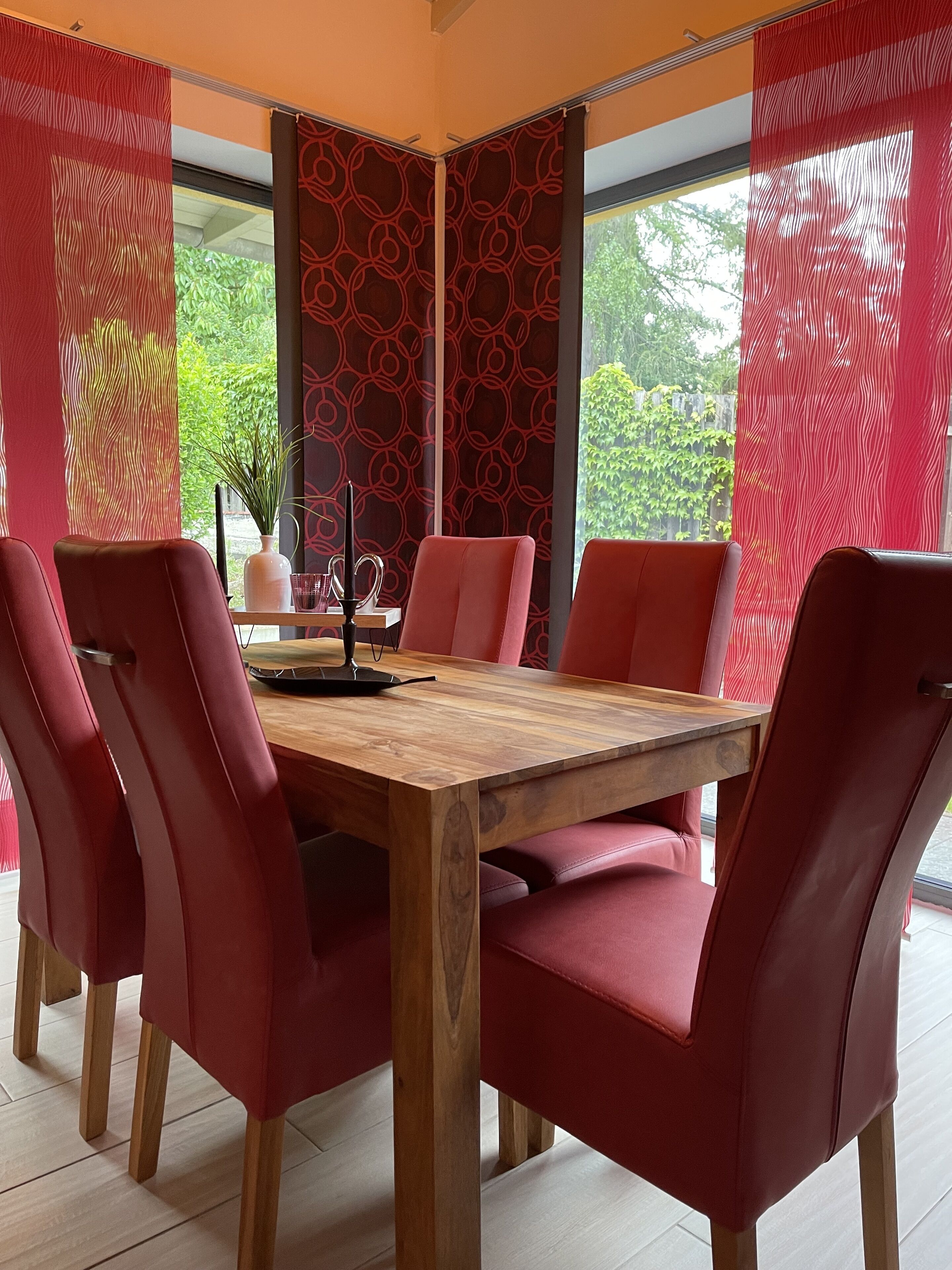 A dining area with a wooden table and red upholstered chairs, surrounded by patterned curtains and a view of greenery outside.