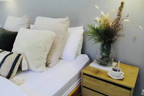 A neatly made bed with white bedding and a fluffy white throw blanket. A small wooden nightstand with a glass vase of dried flowers and a white cup with a spoon is beside the bed.