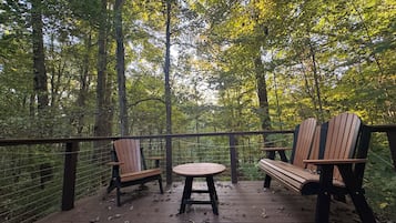 Een houten terras met twee Adirondack-stoelen en een kleine ronde tafel, omgeven door bebossing.