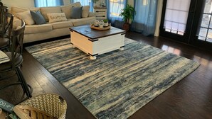 Un salón con sofá, una mesa de centro y un tapete grande. Hay ventanas con cortinas azules y una puerta con persianas.