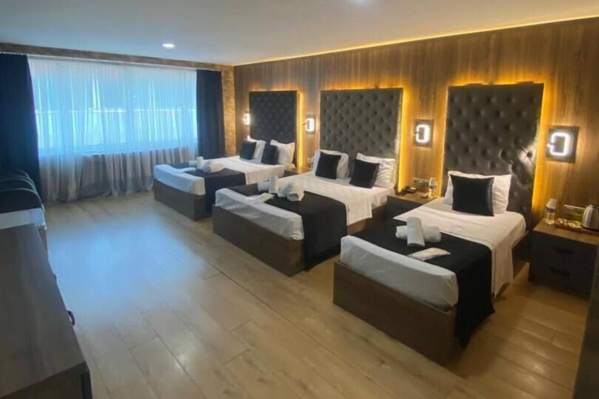 Habitación de hotel con cuatro camas alineadas, suelo de madera y un ventanal con cortinas.