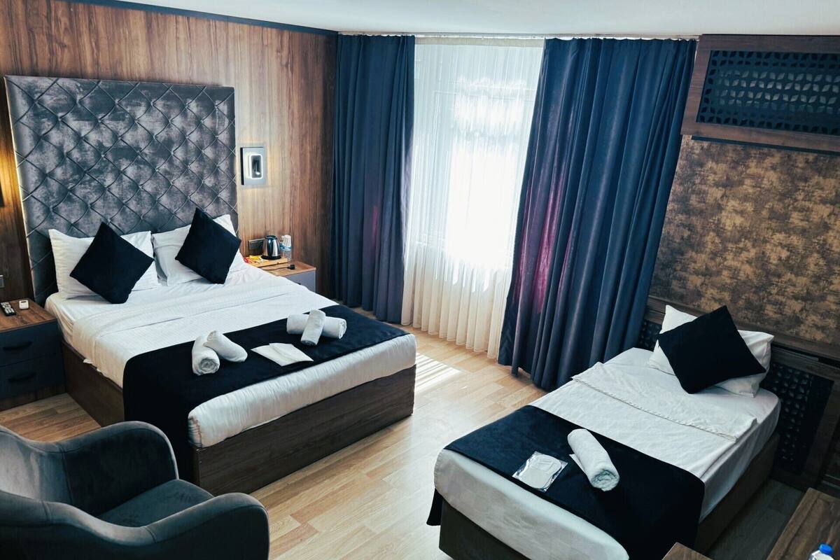 Habitación de hotel con dos camas, cabecera de madera, mesita de noche, una silla y un ventanal con cortinas.