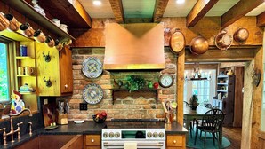 A rustic kitchen with a stone wall, copper cookware, wooden cabinets, and a dining area with a wooden table and chairs.