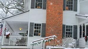 A two-story house with a prominent brick chimney, white siding, and a porch with a railing.
