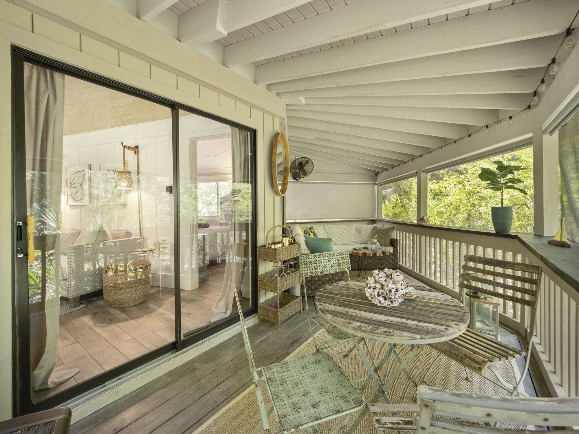 A covered patio with a round table and wicker chairs, a sliding glass door leading to an interior space, and a potted plant on the railing.