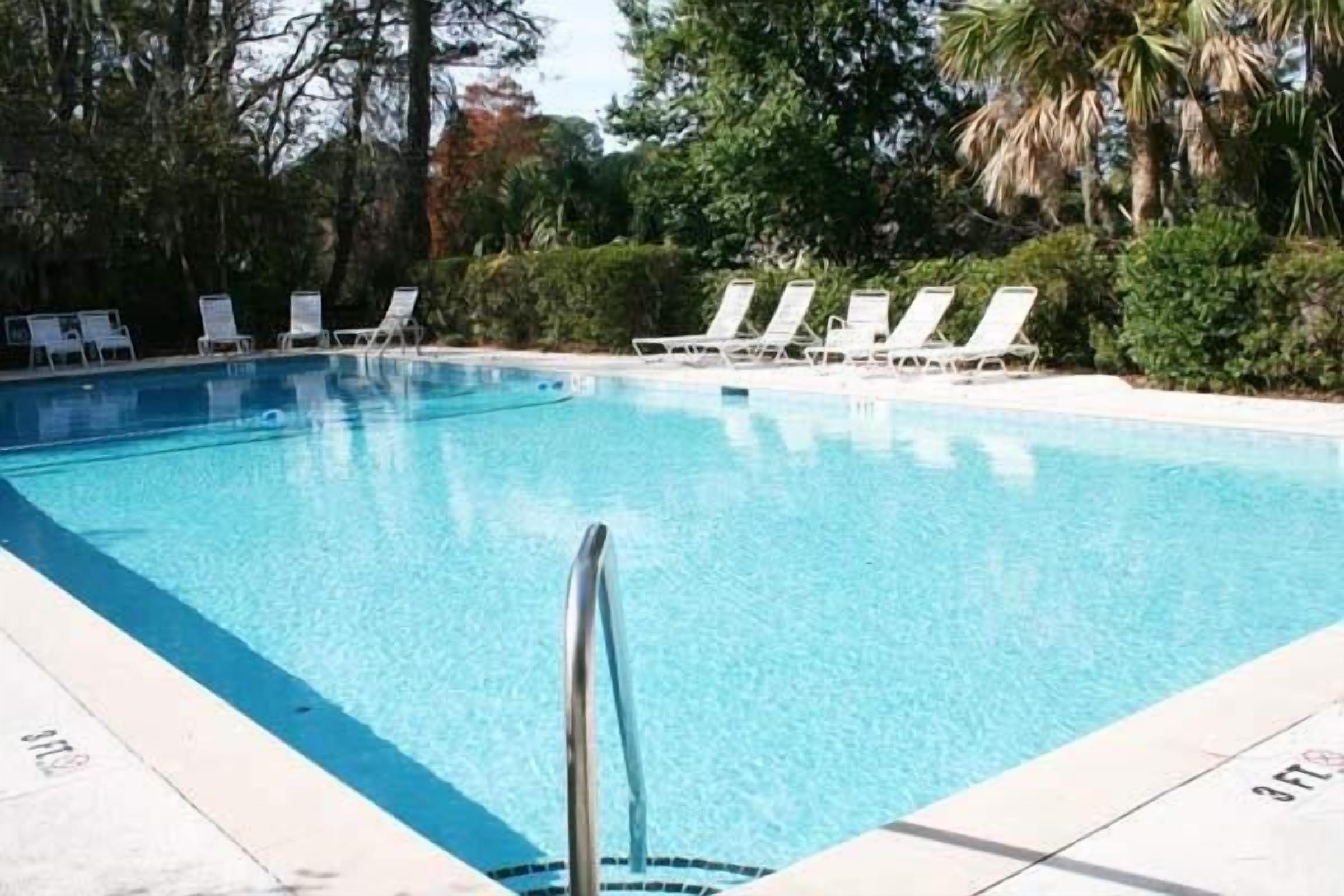 A swimming pool with clear blue water, surrounded by white lounge chairs and lush greenery.