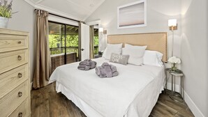 A bedroom with a large bed, a dresser, a nightstand, a lamp, and a view of greenery outside.