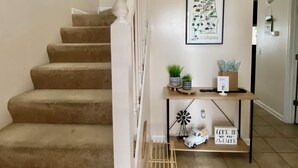 Un escalier dont les marches sont recouvertes d’une moquette beige, avec des rampes blanches et une étagère fixée au mur, ornée d’objets décoratifs et d’une carte encadrée.