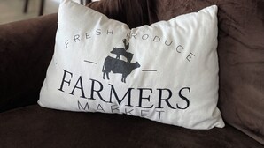 Un salon avec un canapé marron, un coussin blanc orné du texte « MARCHÉ DES PRODUCTEURS DE PRODUITS FRAIS », et une cuisine en arrière-plan.