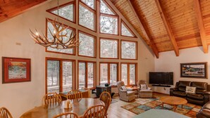 A spacious living area with a high ceiling, large windows, a dining table, and a fireplace.