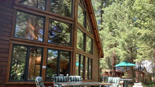 A wooden cabin with large windows, a dining table with striped cushions, and a patio area.