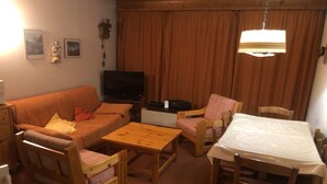 Ein gemütliches Wohnzimmer mit einer Couch, zwei Sesseln, einem Holztisch, einem Fernseher und einem Esstisch mit Stühlen.