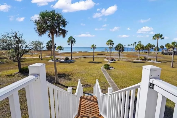 A view from a balcony with a wooden deck leading to a grassy area with palm trees and a beach in the background.