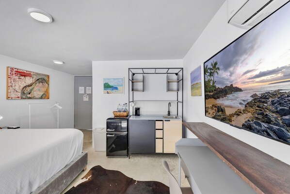 Una habitación de hotel moderna con cama, kitchenette, televisor grande y un cuadro con escena de playa.
