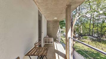 A balcony with a wooden table and bench, a small white cabinet, and a view of a park with trees and grass.