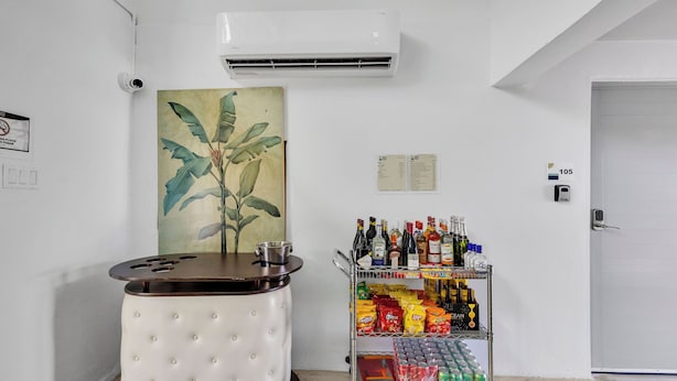 Habitación con aire acondicionado blanco, un cuadro de una planta, un minibar con aperitivos y bebidas, y una puerta con el símbolo de número.
